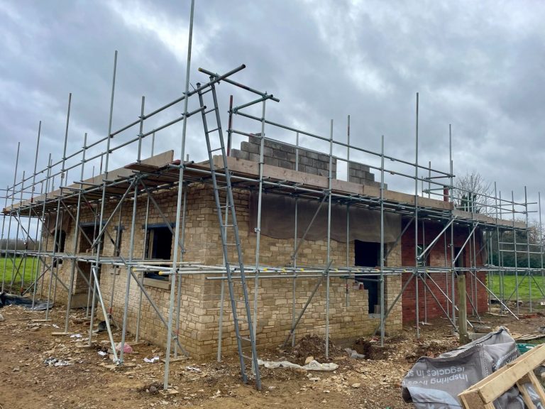 Building under construction with scaffolding, surrounded by a grassy area and cloudy sky.