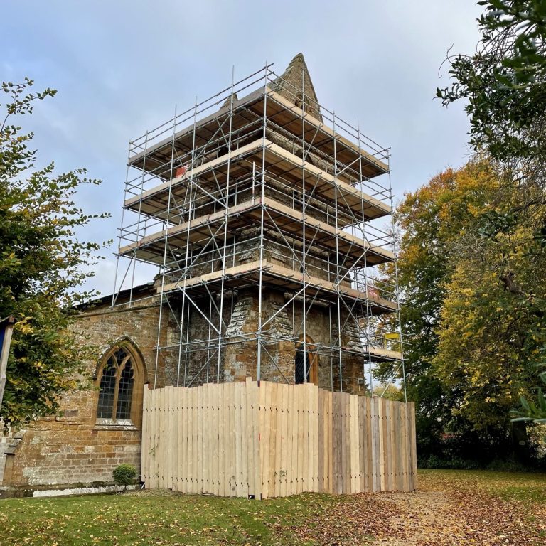 Stone church with scaffolding, surrounded by trees and autumn foliage.