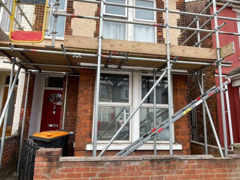 House front with scaffolding set up for renovation, featuring red door and two windows.